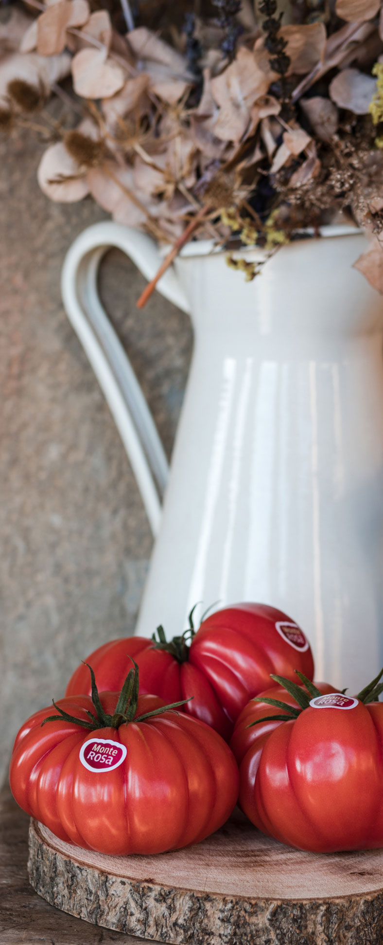 Imagen de 3 Tomates Monterosa apoyados en una rodaja de madera. Al fondo un jarrón con flores preservadas blanco.