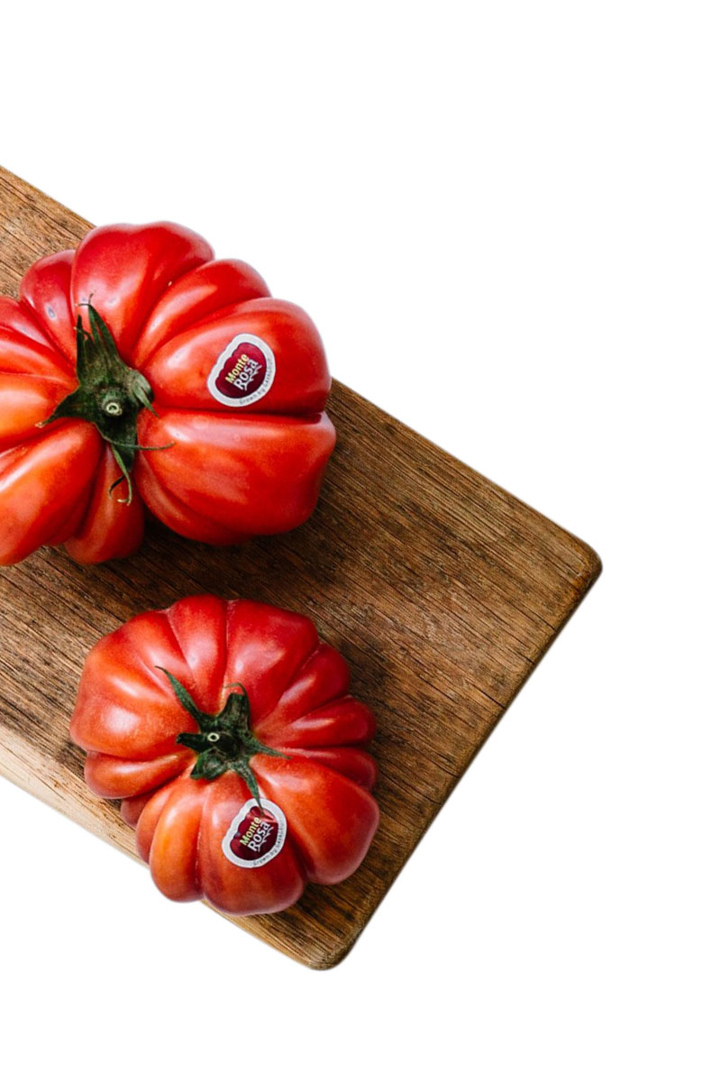 Imagen con fondo blanco. Aparece una tabla de cortar con dos tomates Monterosa vistos desde arriba. Aparece cortada por la izquierda como si saliera de la misma.