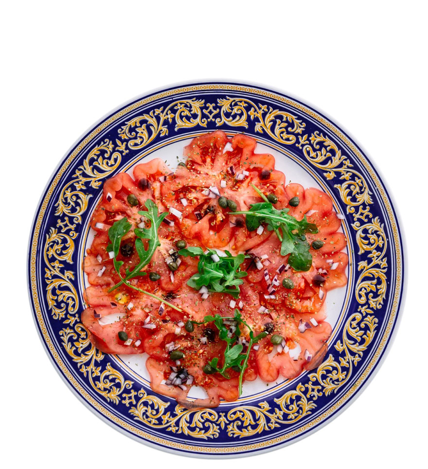 Calorías del tomate, te lo contamos todo. En la imagen un carpaccio, con tomates a rodajas, rúcula, cebolla morada y tápena. Plato visto desde arriba.