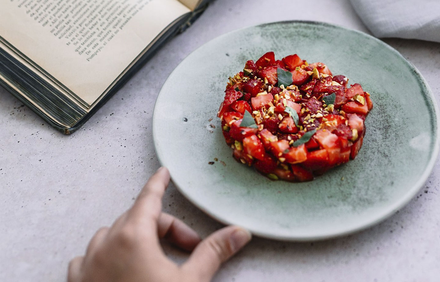 Tartar de tomate Monterosa con fresas. En la imagen la mano de una persona acercándose al plato y un libro de fondo. Tartar en un plato elegante.