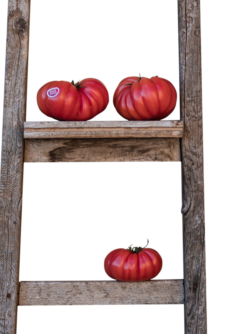 Imagen de una escalera apoyada en una pared blanca con tres tomates posados en el ultimo y penúltimo escalón que es el que se ve.