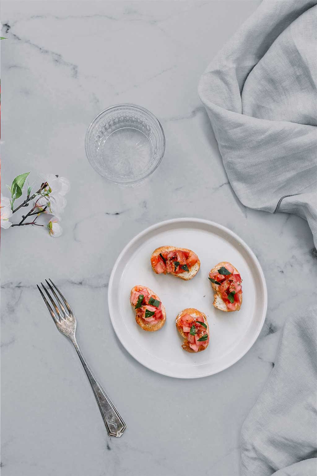 Receta de bocados con Tomate Monterosa y unas hojas de menta a modo de decoración. En la imagen sobre un plato blanco. Tambien se observa un vaso, un tenedor y una rama con flores de azahar. Un trapo de cocina elegante sobre una encimera de mármol.