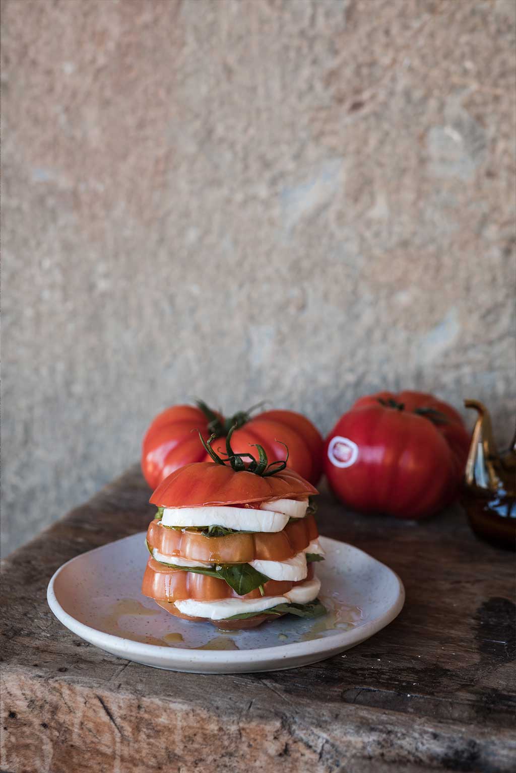 Receta de carpaccio Monterosa . En la imagen un tomate cortado a laminas y entre ellas, rúcula y queso. Aliñado con aceite de oliva. Como si de una torreo se tratara. Presentado sobre un plato en una mesa de diseño rústico. Dos tomates enteros al fondo.