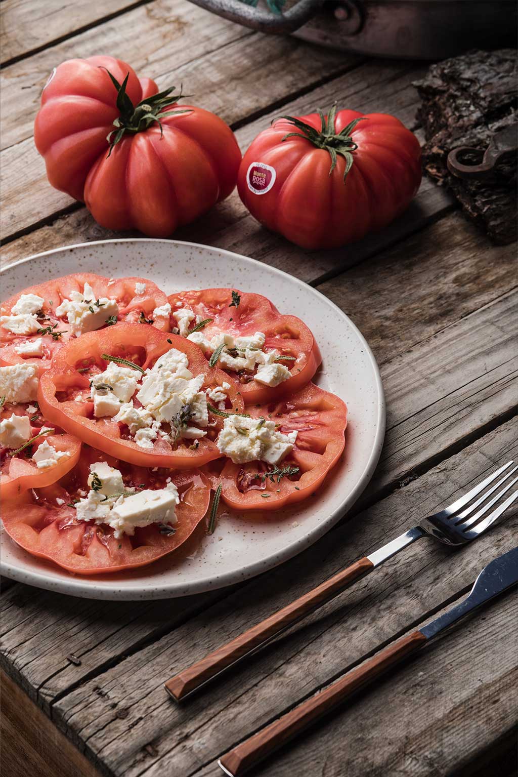 Recetas con tomates monterosa. Carpaccio con queso de cabra, hierbas y Aove. Sobre una mesa rústica con cuchilloy tenedor a la derecha y dos tomates enteros al fondo.