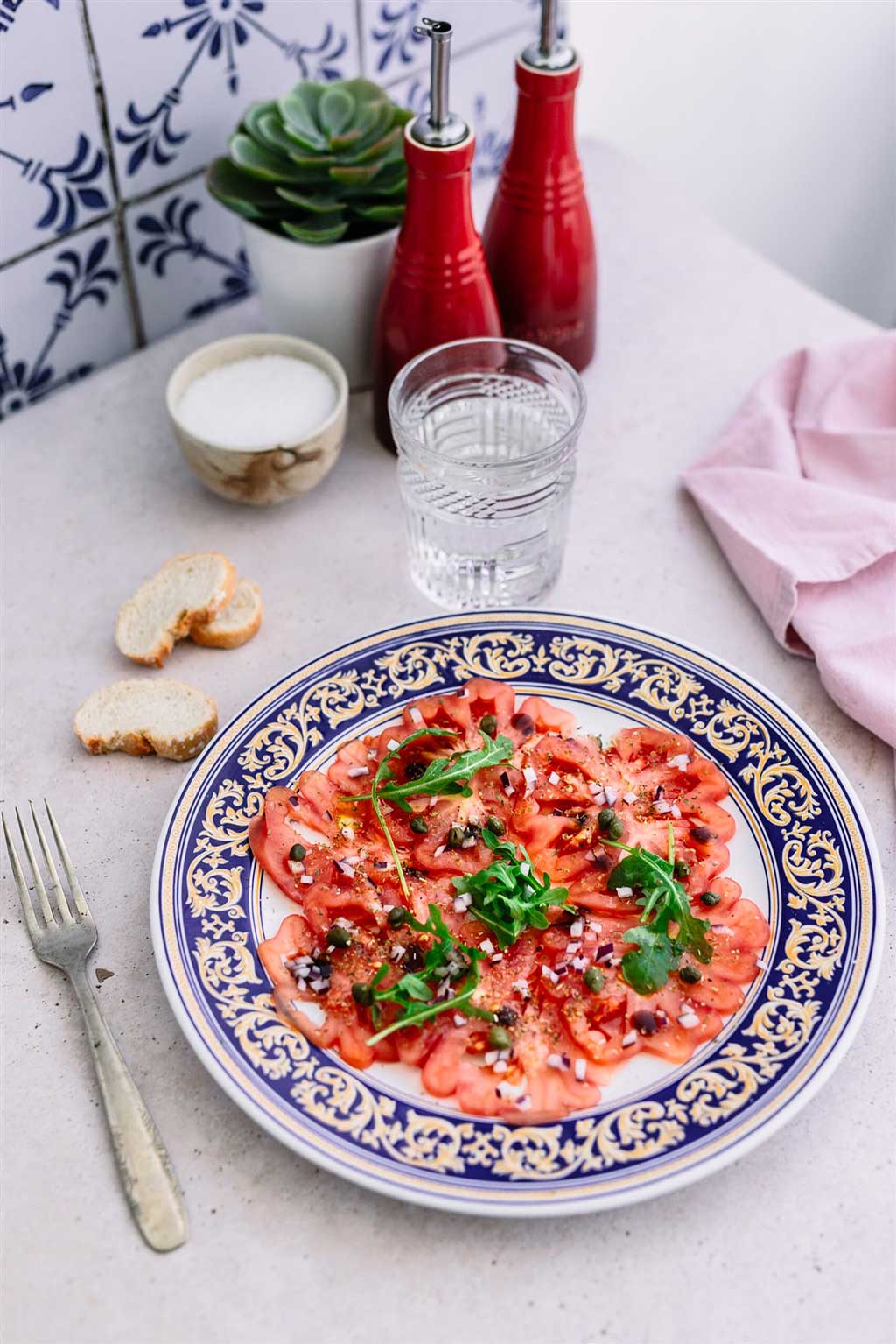 En la imagen un carpaccio, con tomates a rodajas, rúcula, cebolla morada y tápena. En la mesa listo para comer se ve un tenedor a la izquierda, unas totadas arriba junto a un vaso de agua. Un recipiente con sal y otros dos con aceite de oliva y vinagre. Y una plantita de decoración. Foto usada para explicar la receta que se ve.