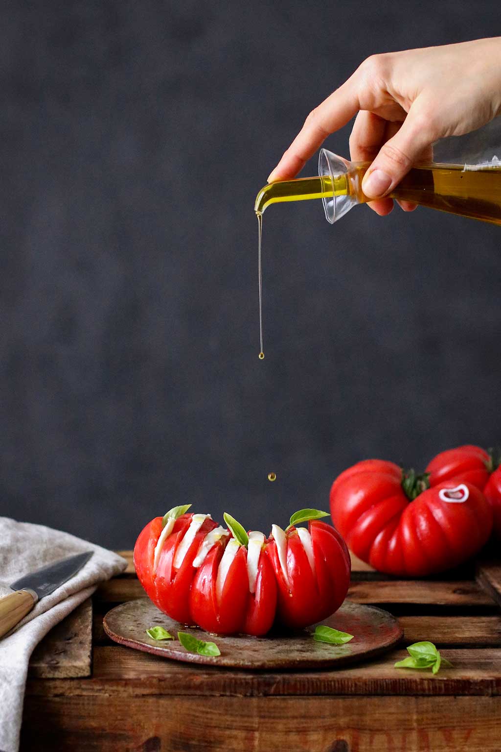 Receta Monterosa Capresse. En la imagen en una mesa rústica sobre un plato de barro un tomate con cortes con mozzarela entre ellos. Una forma original de mostrar esta receta. La mano de una mujer echa aceite de oliva sobre el. Al fondo un tomate entero y un cuchillo.