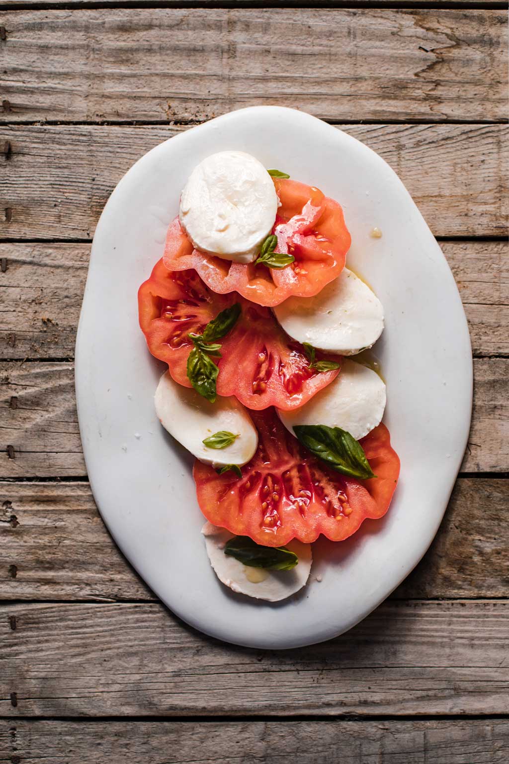 Receta de ensalada con Tomate Monterosa, Queso mozzarella y unas hojas de albahaca a modo de decoración. En la imagen sobre un plato blanco alargado sobre una mesa de madera.