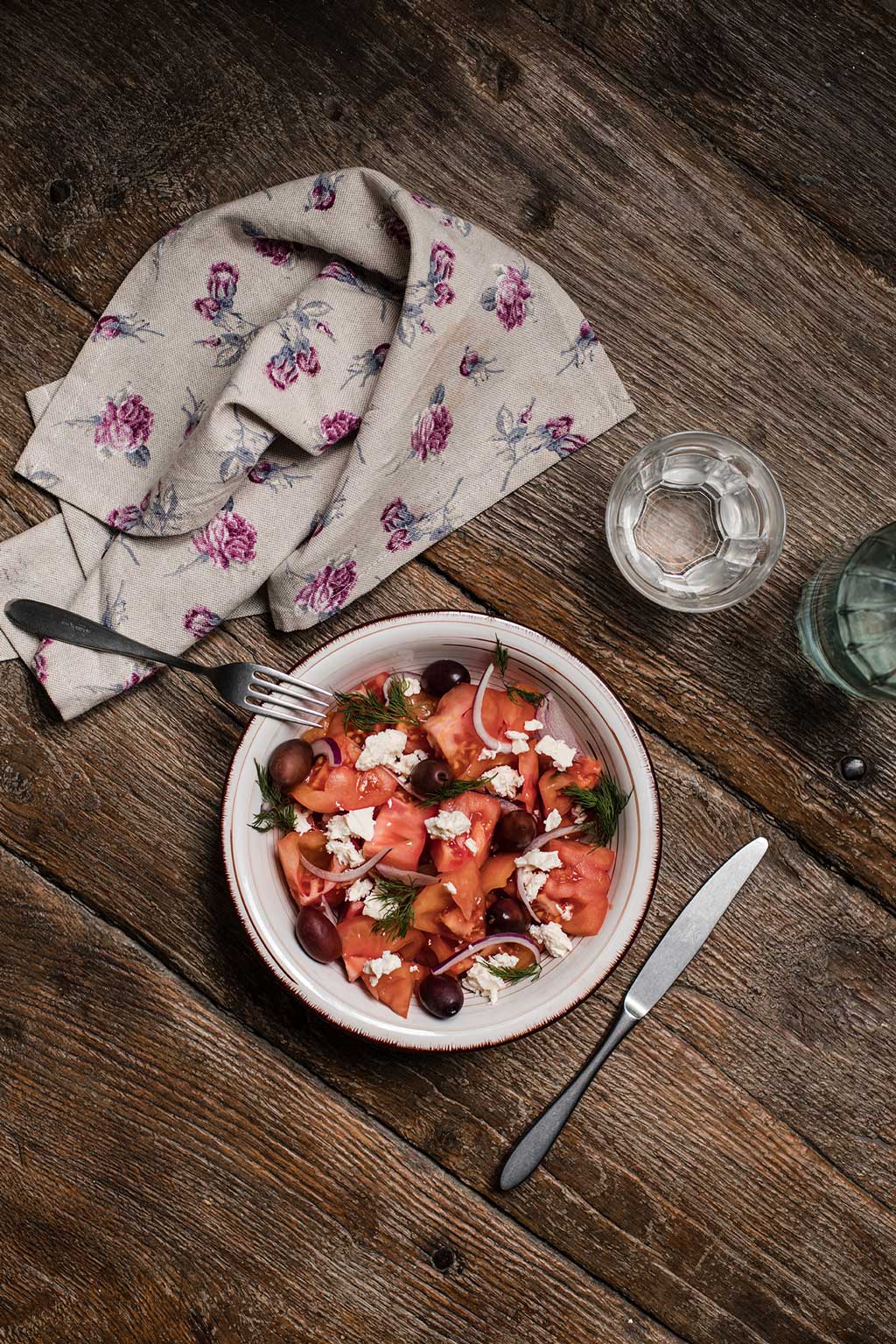 Receta de ensalada con Tomate Monterosa, cebolla morada, queso feta y olivas, sobre una mesa rústica preparada para comer se ve el tenedor, el vaso de agua y la servilleta. Foto echa desde arriba