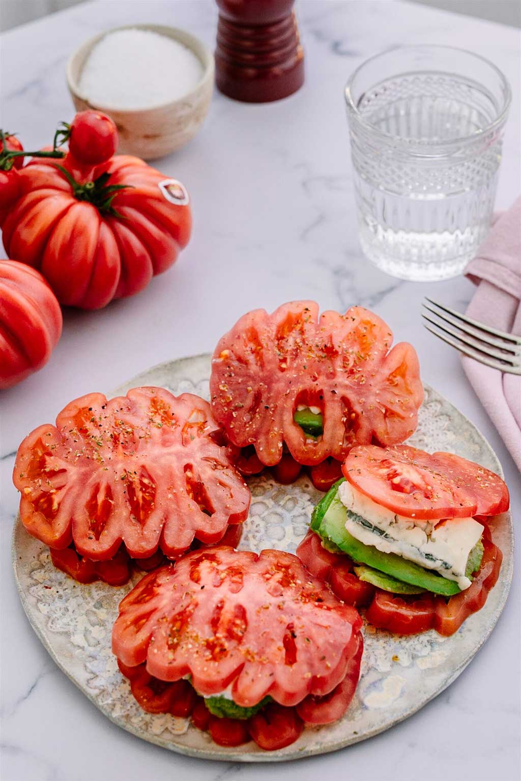 Imagen de una ensalada Con tomates Monterosa montados como si fueran sandwich con aguacate y queso azul dentro. Sobre un plato. A la derecha un tenedor y dos tomates enteros, un vaso de agua y sal. Esta imagen se usa para indicar lla receta de esta ensalada.