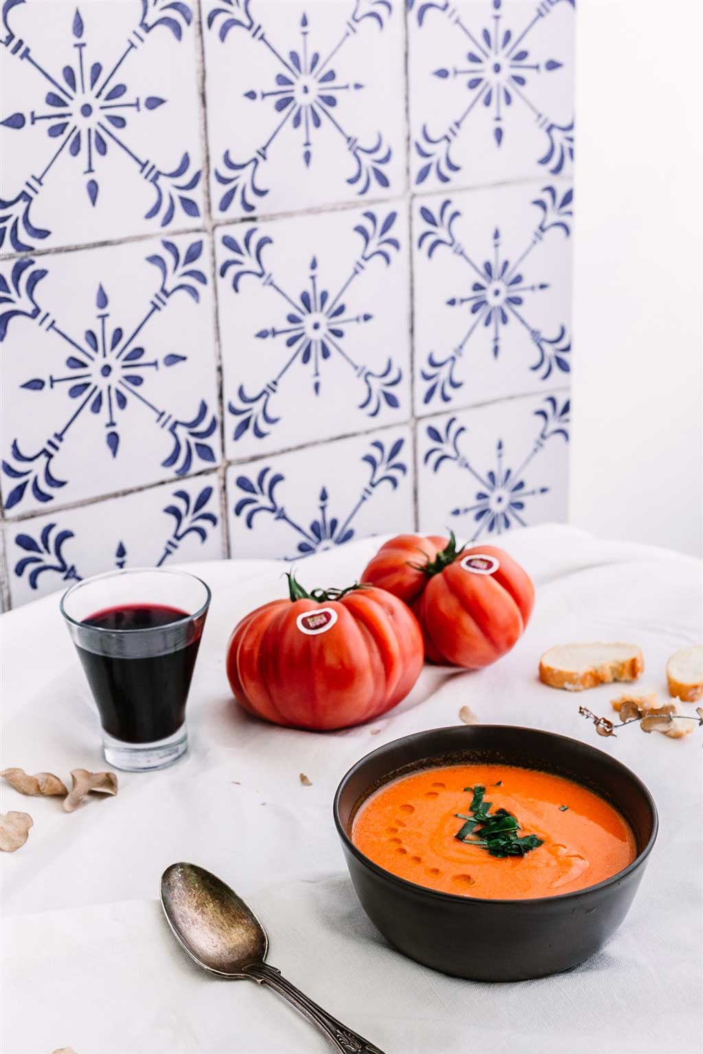 Recetas con tomate Monterosa Gazpacho Rosa. En la imagen aparece un bol de gazpacho de color naranja. Se ven unas gotas de aceite con el que se ha aliñado y unos trocitos de remolacha. A la izquierda una cuchara para tomarlo. A la derecha unas tostas para acompañarlo. Arriba un vaso de vino y don tomates enteros. La pared de fondo en azulejo blanco con dibujos azules antiguos. Foto principal de esta receta.