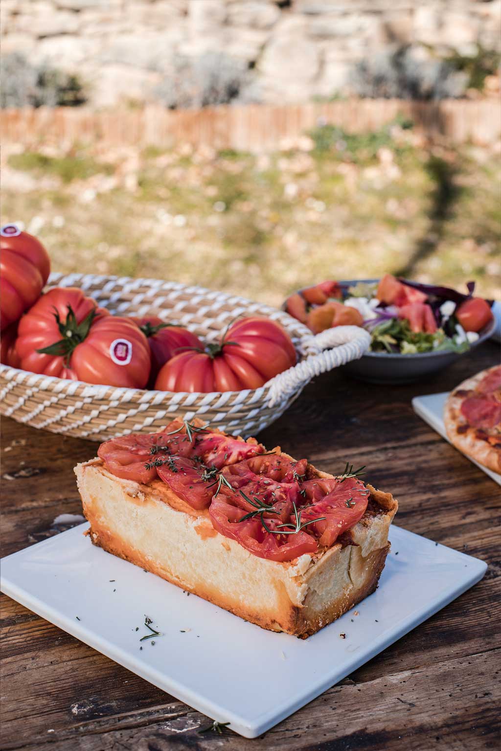 Receta de Hojaldre con Tomate Monterosa. En la imagen sobre un plato blanco cuadrado. También se observa una ensalada y un cesto con tomates y el filo de una pizza. La foto está echada en el exterior.
