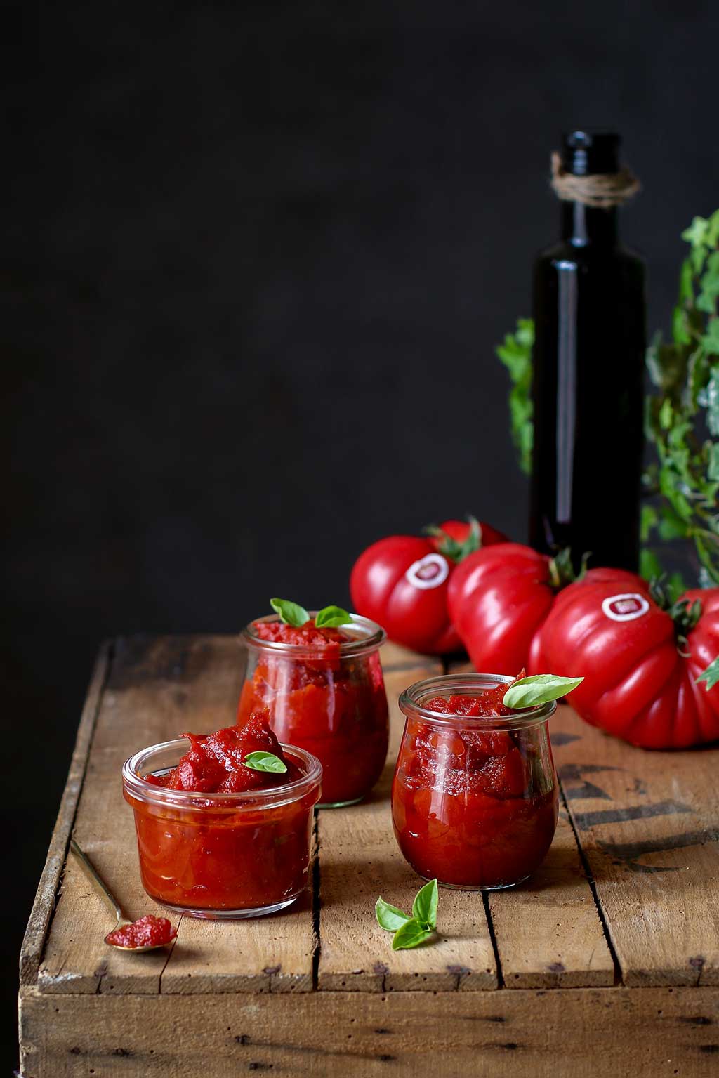 Receta de passata con Tomate Monterosa un plato típico italiano presentado en tarritos de cristal. En la imagen sobre una mesa de madera rustica. Tambiense ven tres tomates enteros un poco de sal y un aceite de oliva y una cuchara para comerlo.