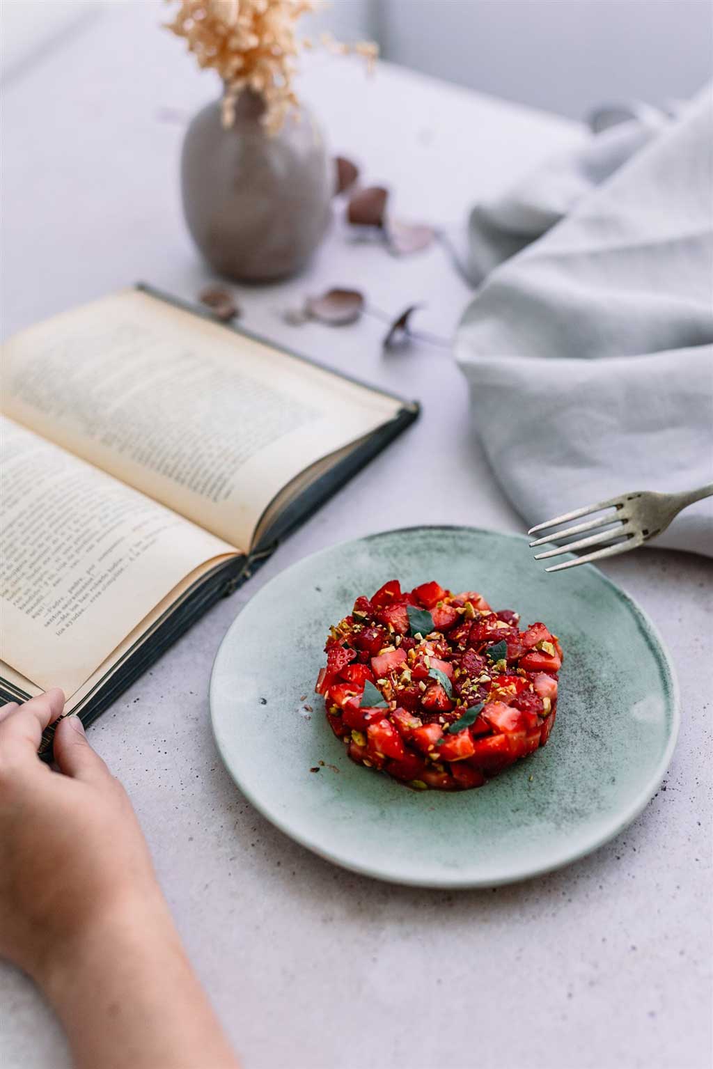 Tartar de tomate Monterosa con fresas. en la imagen una persona lo disfruta mientras lee un libro. Tartar en u plato elegante.