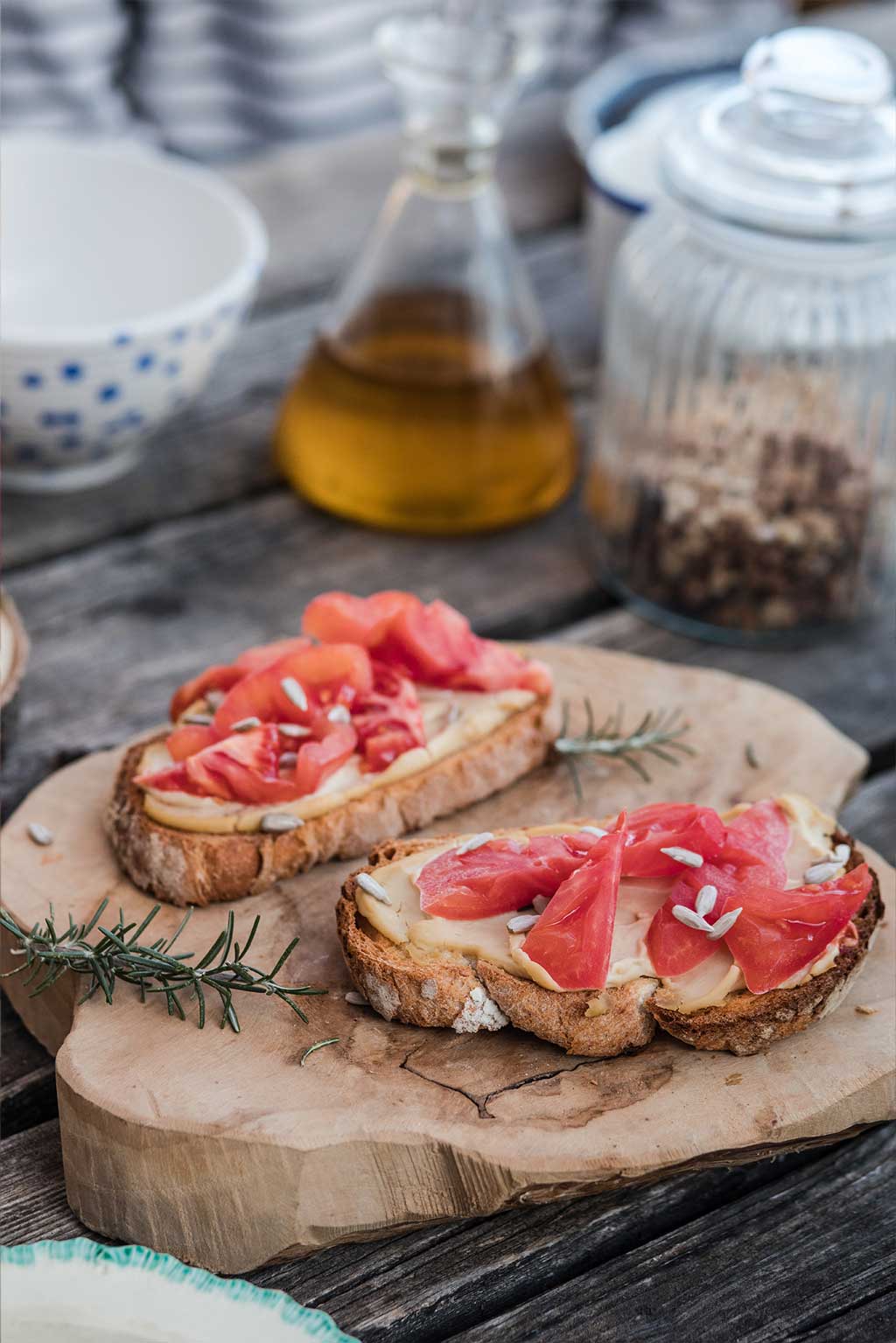 Receta de tostadas de pan rústico con Tomate Monterosa, humus y frutos secos. En la imagen sobre una rodaja de madera con ramas de romero alrededor. También se observa un bote con los frutos secos y un recipiente con aceite de oliva.