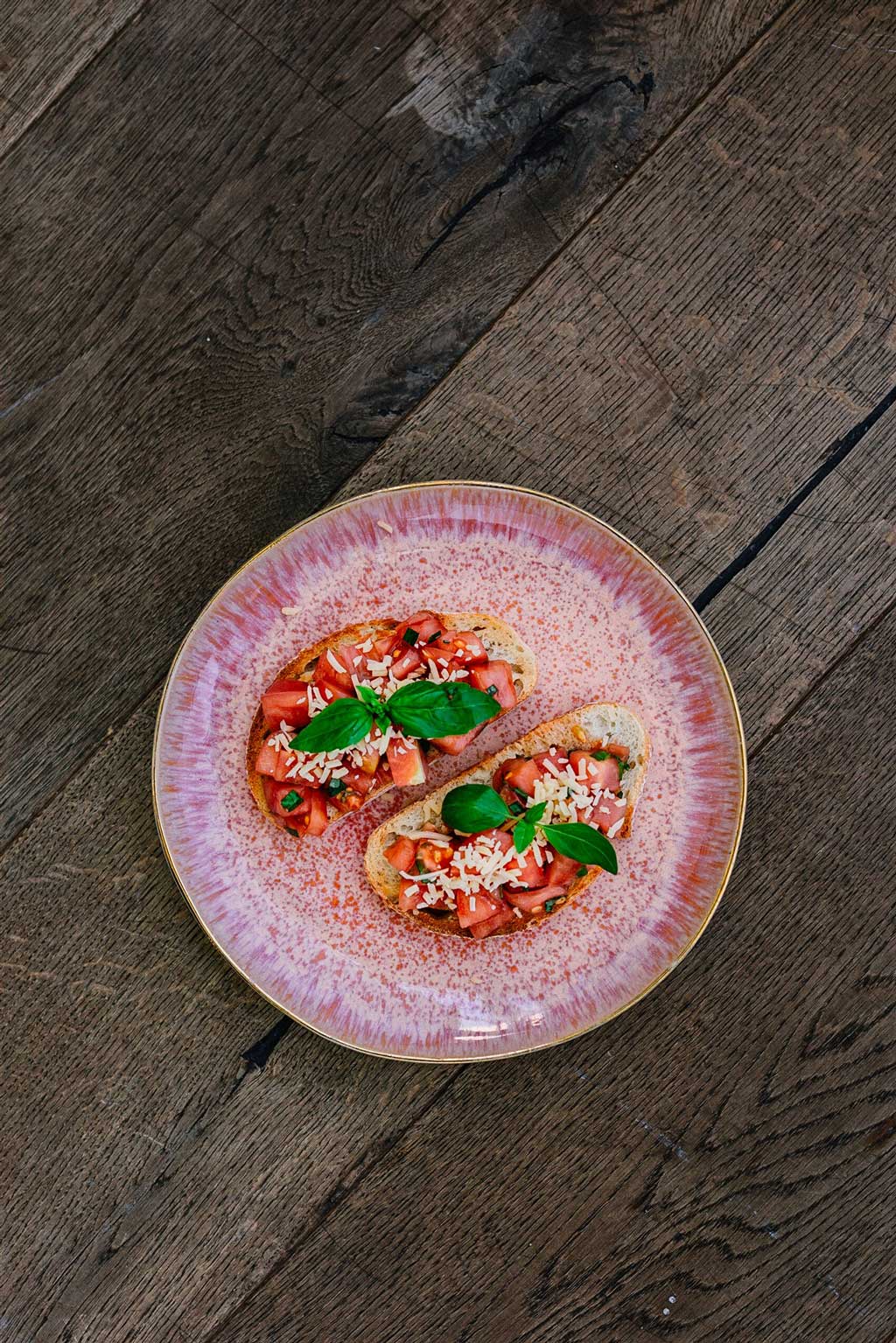 Receta de tostadas de pan rústico con Tomate Monterosa, Queso y unas hojas de albahaca a modo de decoración. En la imagen sobre un plato rosa sobre una mesa de madera.