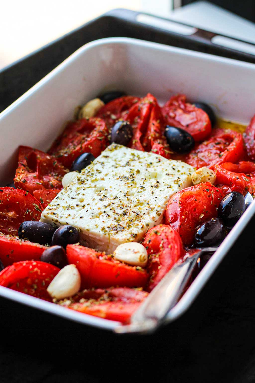 Receta de queso feta al horno con aceitunas y tomate monterosa. By Laura García. En la imagen sobre un bol cuadrado encima de la bandeja del horno con cuchara dentro para servir.