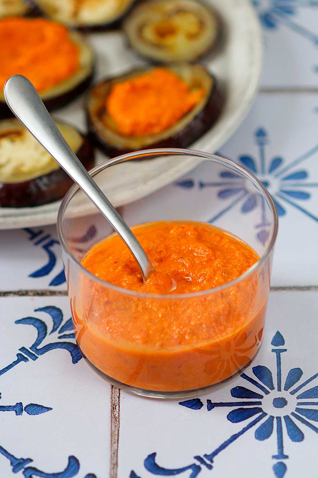 Receta de Salsa Romesco con tomate Monterosa. Presentado vaso de cristal redondo y una cuchara dentro para tomarlo. Sobre una encimera de azulejo blanco y azul. De fonde calabacines rellenos de la misma. By Marina Ribas.